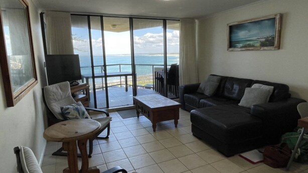 A living room with a sofa, a TV, a table, and a balcony with a view of the sea.