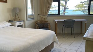 A bedroom with a bed, a desk, a chair, a lamp, and a window with a view of the sea.