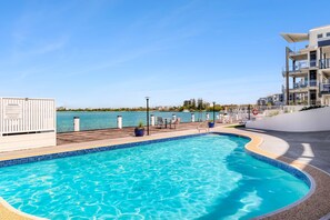 A swimming pool with a view of the sea, a deck area with chairs, and a building with balconies. - Pumicestone Chase U8 Jetty, Pool & Views (Caloundra)