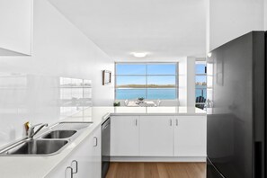 A modern kitchen with a large window overlooking a body of water, white cabinetry, and a stainless steel sink. - Pumicestone Chase U8 Jetty, Pool & Views (Caloundra)