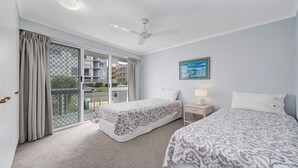 A hotel room with a balcony, two beds, a ceiling fan, a nightstand with a lamp, and a painting on the wall.