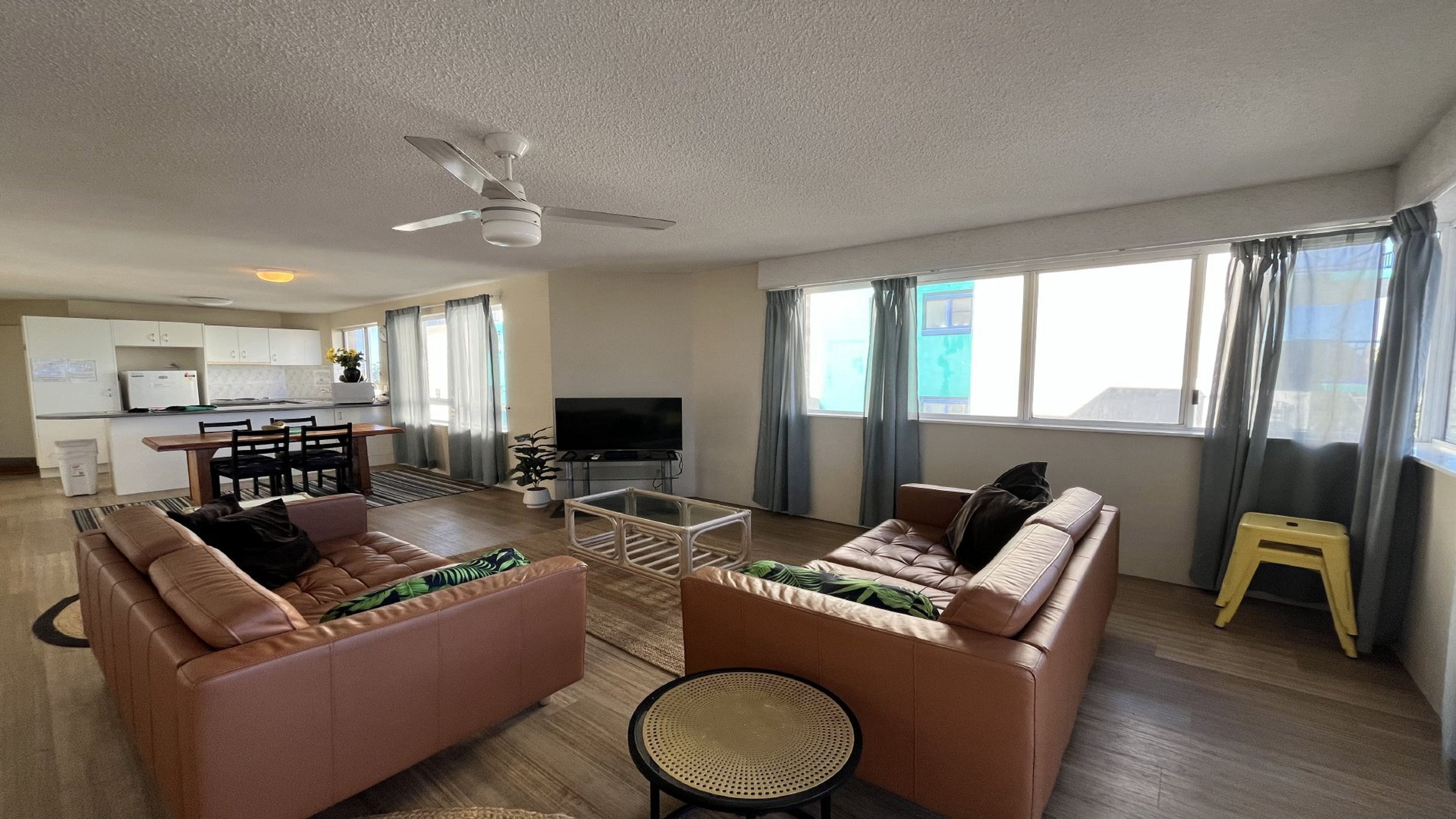 A living room with two sofas, a coffee table, a TV, and a ceiling fan. There is a kitchen in the background with a dining table and chairs.