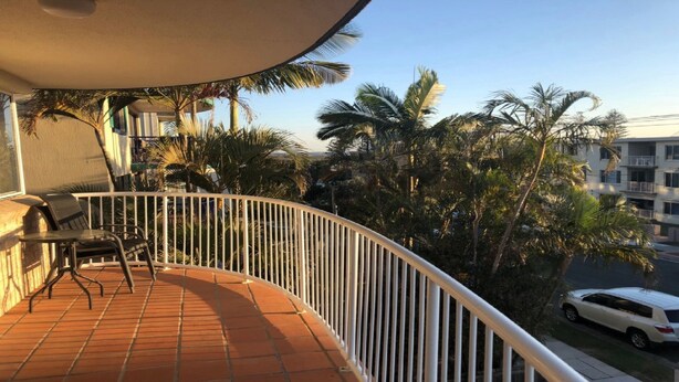 Un balcón con suelo de baldosas, baranda blanca y vista a palmeras y un edificio.