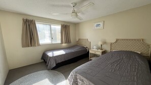 Una camera d'albergo con due letti, una ventola a soffitto, una finestra con tende, un comodino con una lampada e un quadro appeso al muro.
