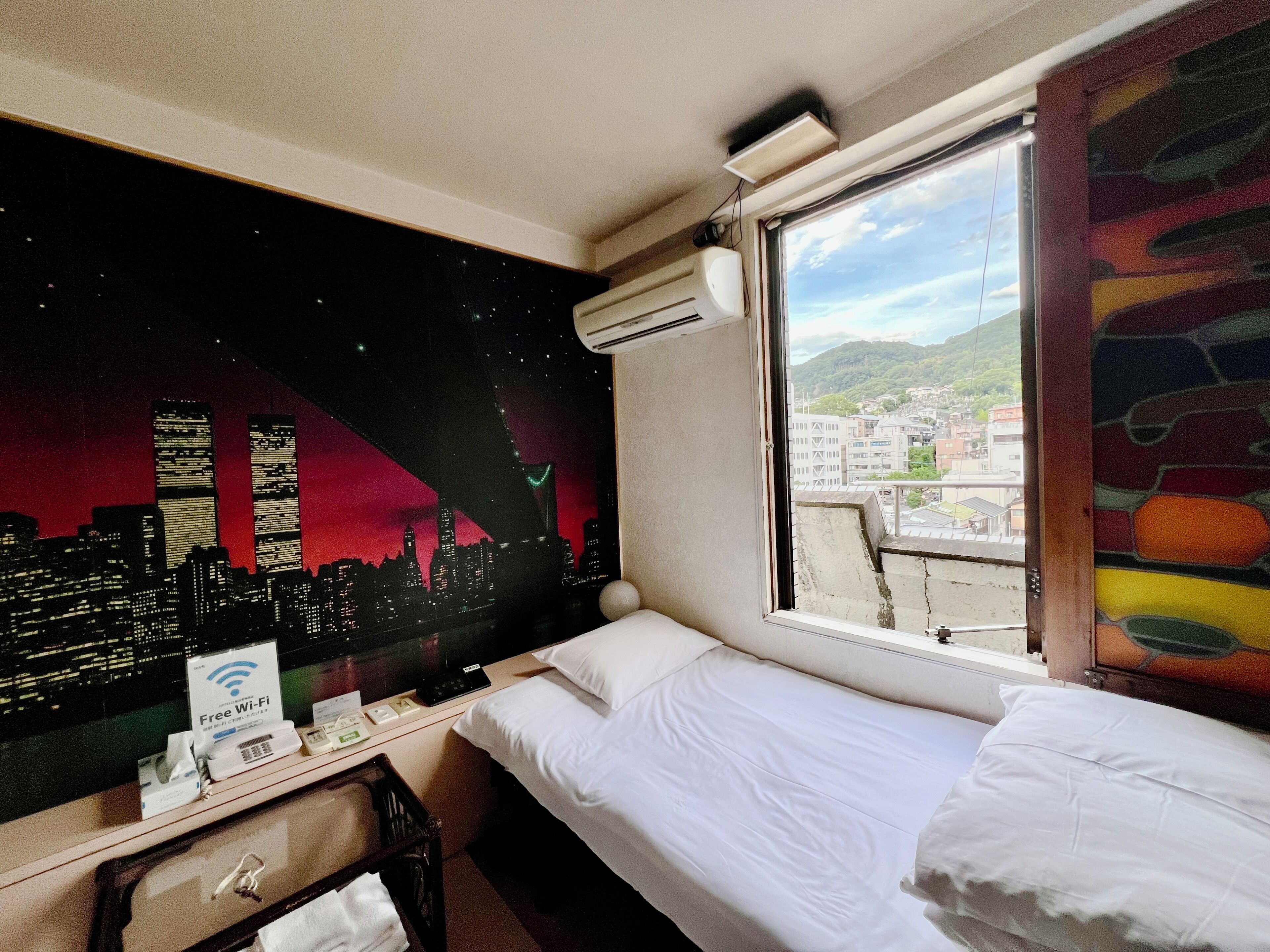 A hotel room with a bed, a desk with a computer and a sign indicating free Wi-Fi, a window with a view of a cityscape and mountains, and a wall with a cityscape mural.