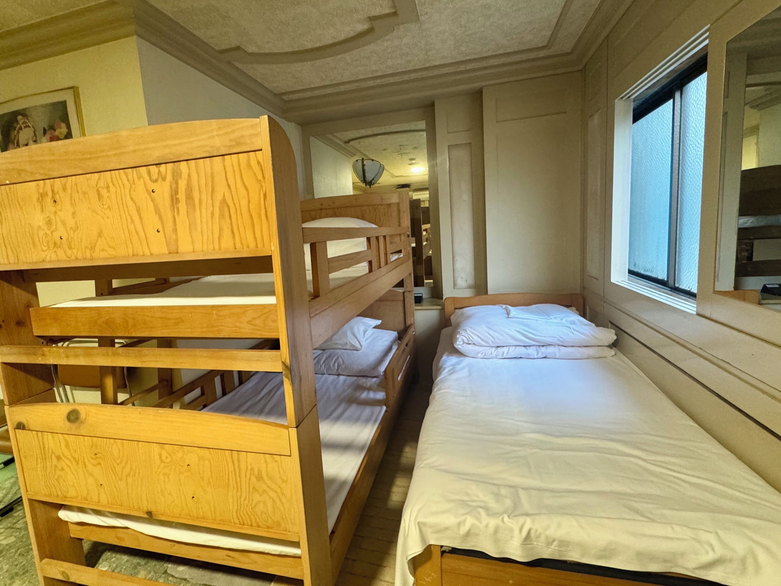 Une chambre avec un lit superposé et une fenêtre, des lits superposés en bois et un lit simple avec une literie blanche.