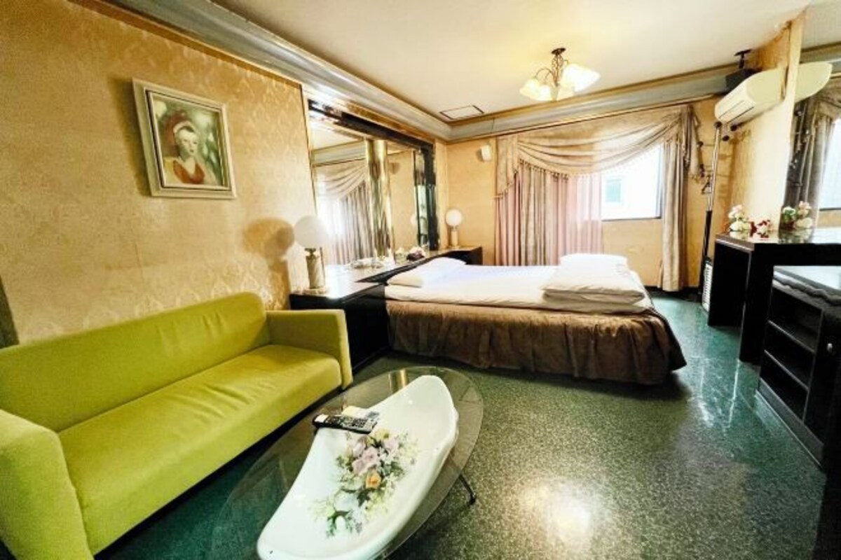 A hotel room with a bed, a green sofa, a glass table with a floral design, and a large mirror.