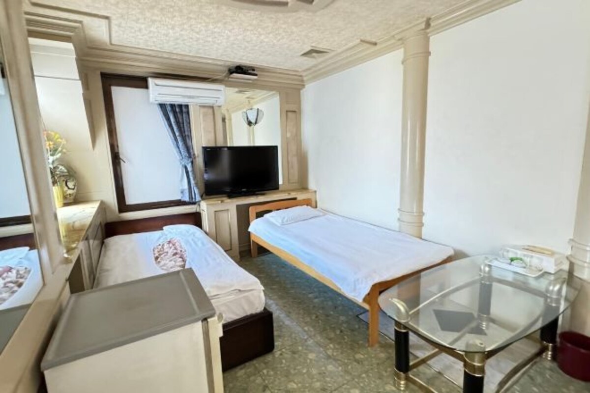 A hotel room with two single beds, a television, a small table, and a window with curtains.