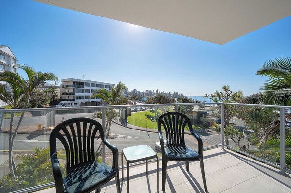 A balcony with two chairs and a small table, overlooking a street with buildings and palm trees.