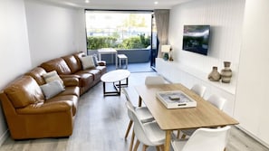 A modern living room with a brown leather sofa, a wooden dining table, white chairs, a TV mounted on the wall, and a view of greenery outside.