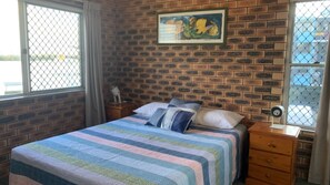 A room with a brick wall, a bed with a striped blanket, a nightstand, a wooden dresser, and a window with a view. - Tingira Sea 2 Steps To Bulcock Beach & Shops (Caloundra)