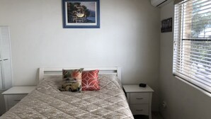 Een slaapkamer met een bed, een nachtkastje, een raam met jaloezieën en een ingelijste foto aan de muur.