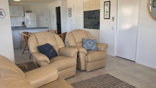 Una sala de estar con dos sillones color beige, un comedor con sillas de mimbre y una cocina con gabinetes blancos.
