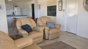 A living room with two beige armchairs, a dining area with wicker chairs, and a kitchen with white cabinets.