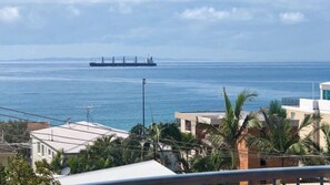 Vista costera con un gran buque de carga en la distancia, edificios con antenas parabólicas y palmeras.