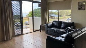 A living room with a black leather sofa, a glass coffee table, and a view through a sliding glass door to an outdoor area with a car and a building.