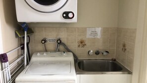 A compact laundry area with a washing machine, a sink, and a sign instructing to remove clothes before using the dryer.