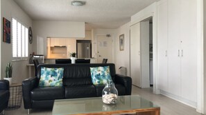 Een moderne woonkamer met een zwarte leren bank, een glazen salontafel en een keuken op de achtergrond.