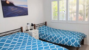 Een slaapkamer met twee bedden, een nachtkastje, een bureau en een raam met jaloezieën.