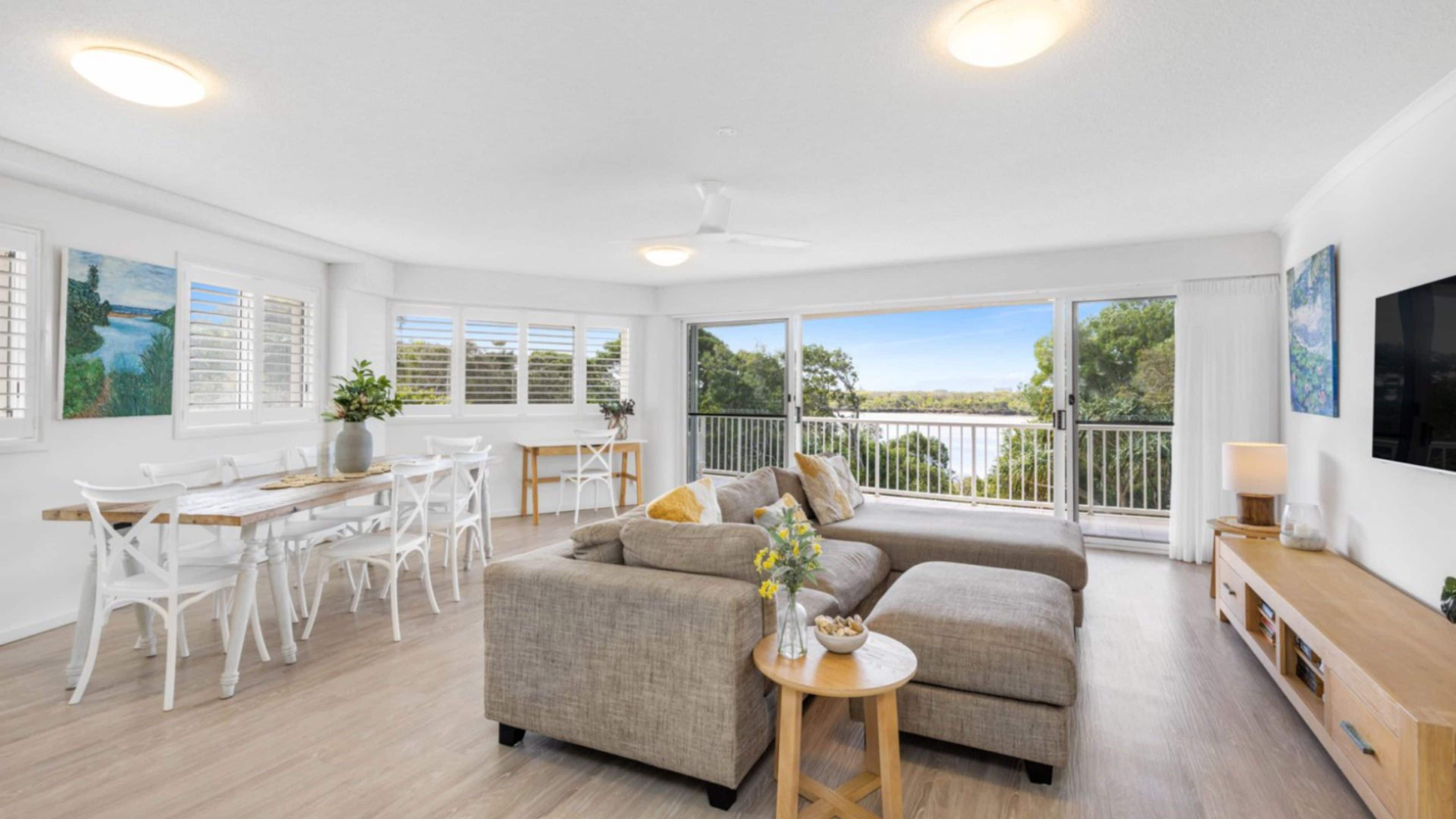 A spacious living area with a large sofa, dining table, and chairs, a balcony with a view, and a flat-screen TV mounted on the wall.
