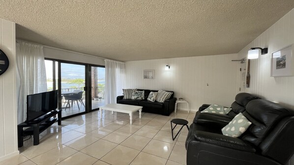 A living room with a black leather sofa, a white coffee table, a black TV stand, and a sliding glass door leading to a balcony. - Balamara U3 Water Views, Walk to Bulcock Beach (Caloundra)