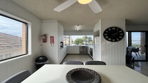 A modern kitchen with a large window, a dining area with a white table, and a black clock on the wall. - Balamara U3 Water Views, Walk to Bulcock Beach (Caloundra)