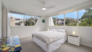 Un dormitorio con cama, mesitas de noche, ventilador de techo y ventanas amplias.