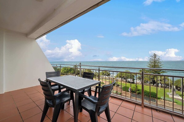 A balcony with a table and chairs, overlooking a coastal view with a clear sky.