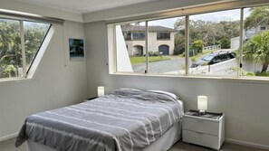 Quarto com cama, mesas de cabeceira e janela com vista para uma casa e rua.