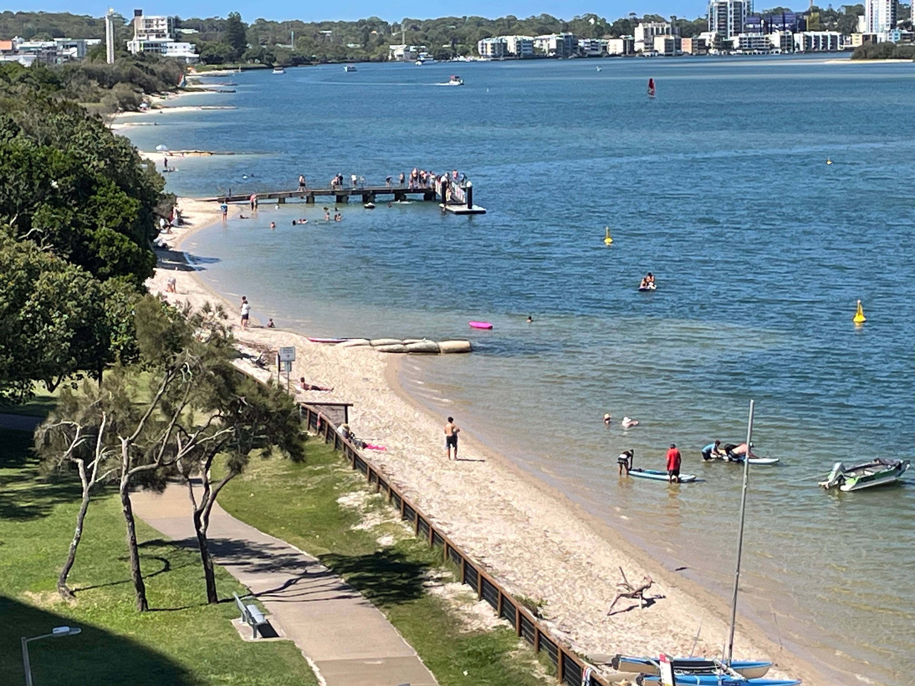 A beach with a wooden pier, clear water, and a city skyline in the background.