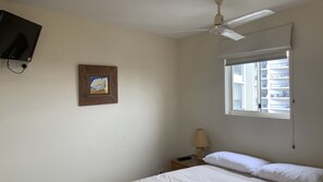 Una camera da letto con un letto, un comodino, una lampada, una ventola a soffitto, una finestra con persiane e un quadro appeso al muro.
