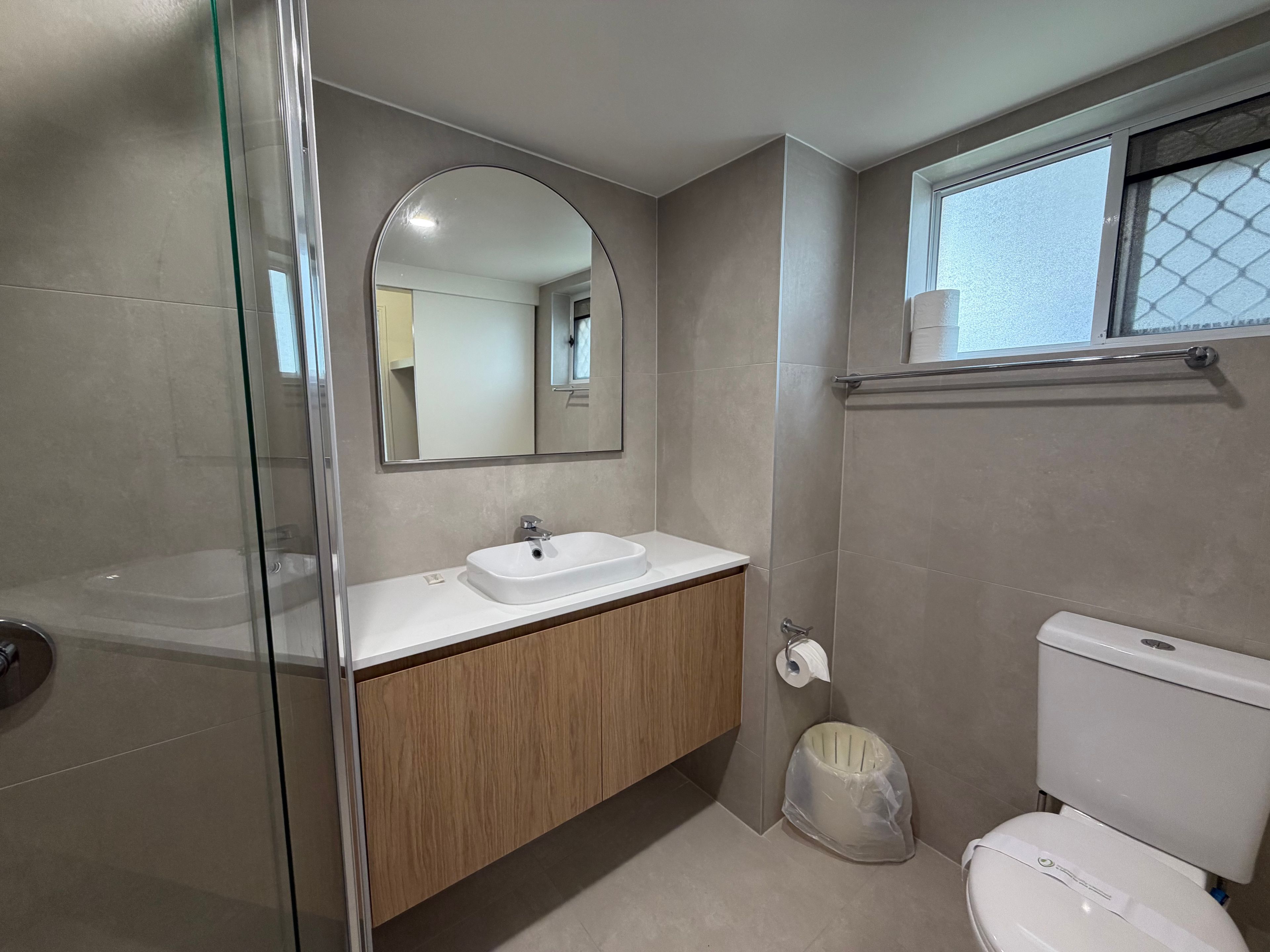 Een badkamer met een douchecabine van glas, een witte wastafel met een spiegel erboven, een toilet en een raam met uitzicht.