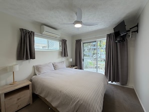 Une chambre à coucher comprenant un lit, une table de chevet, une lampe, un climatiseur, un ventilateur de plafond, un téléviseur fixé au mur et une porte-fenêtre coulissante donnant sur un espace extérieur.