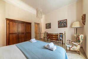 A bedroom with a bed, a wooden wardrobe, a brass bed frame, a floral armchair, and two wall-mounted paintings.