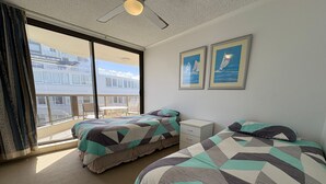 A hotel room with two beds, a small bedside table, a window with a view of a building, and a ceiling fan.