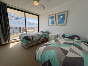 A hotel room with two beds, a small bedside table, a window with a view of a building, and a ceiling fan.