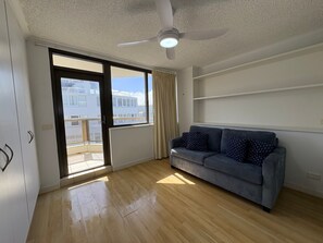 Una habitación con sofá gris, ventilador de techo, puerta corredera de vidrio y vistas a edificios.