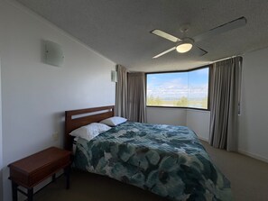 Un dormitorio con cama, mesita de noche, ventilador de techo y ventana con cortinas.