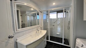 Un baño con tocador blanco, un espejo grande, cabina de ducha con mampara de vidrio y un inodoro.