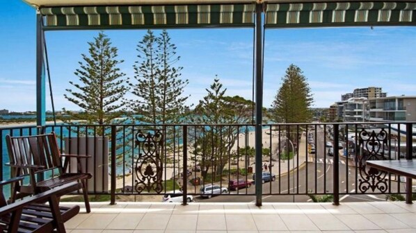 Un balcón con vista a la playa y al paisaje urbano, que incluye una barandilla y una mesa con sillas.