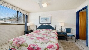 A bedroom with a bed, two bedside tables, a chair, a picture on the wall, and a window with blinds.