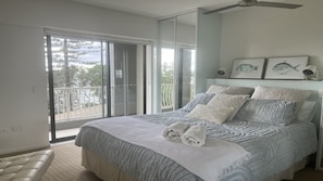 A bedroom with a large bed, a ceiling fan, a balcony with a view of trees, and two framed fish paintings on the wall.