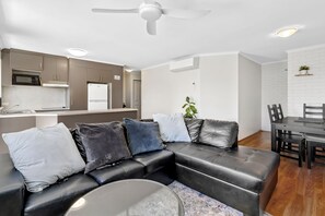 A modern living room with a black leather sofa, a dining table with chairs, and a kitchen area with a refrigerator and microwave.