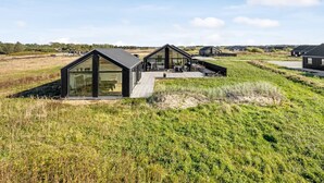 A modern house with large glass windows and a wooden deck, surrounded by grassy fields and other similar houses.