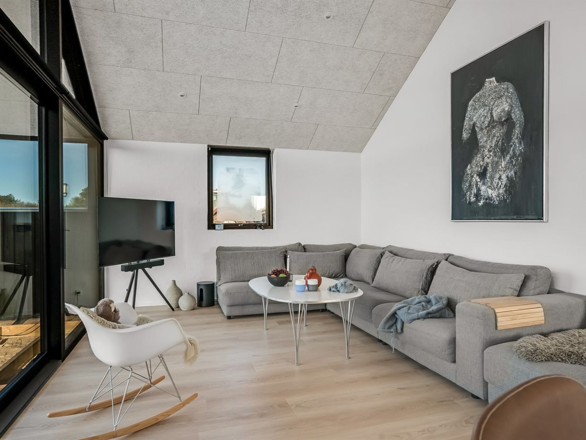 Una sala moderna con un sofá gris, un sillón mecedora blanco, una pequeña mesa redonda y un cuadro abstracto de gran tamaño.