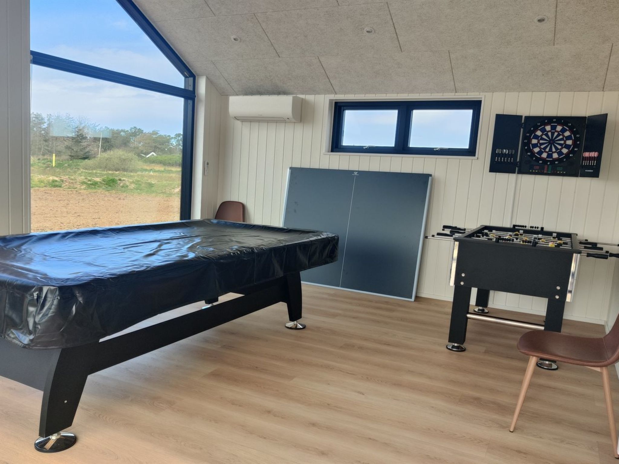 A room with a pool table, foosball table, dartboard, and large windows offering a view of the outdoors.