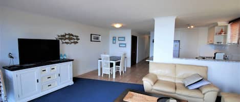 A living room with a flat-screen TV, a dining table with chairs, a beige sofa, and a kitchen area in the background.