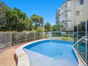 A swimming pool with a white fence, surrounded by a building with balconies and a clear blue sky.