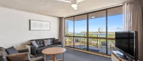 A modern living room with a leather sofa, a wooden coffee table, a flat-screen TV, and a large sliding glass door leading to a balcony with a view of the outdoors.