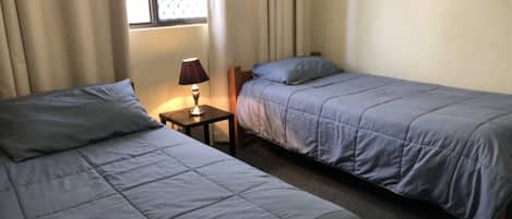 A room with two beds, a nightstand with a lamp, and a window with curtains.